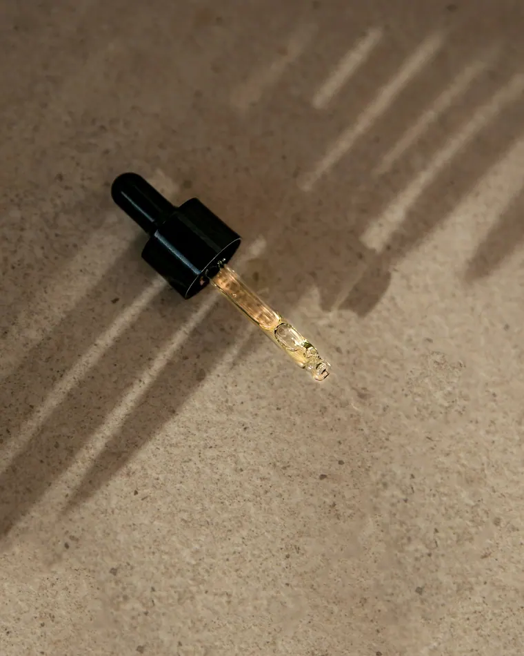 Conta-gotas de vidro com sérum dourado sobre pedra cor de areia, sob sombras de folhas.